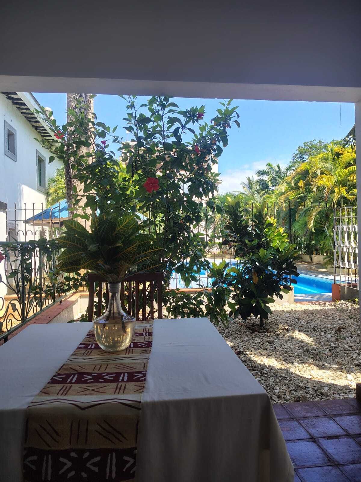 A table with a patterned tablecloth, a vase, and a view of a swimming pool and tropical plants.