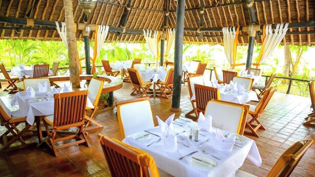 An outdoor dining area with round tables and wooden chairs, covered by a thatched roof. - Mangro Hotel Diani Beach (Ukunda)
