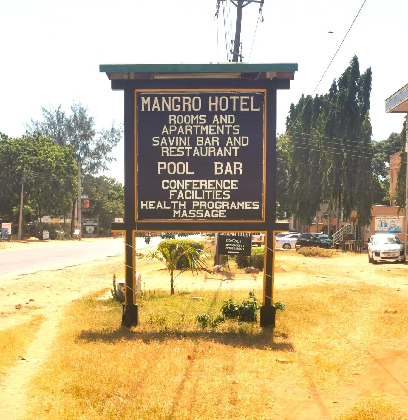 A signboard for Mangro Hotel listing amenities such as rooms, apartments, a bar, restaurant, pool bar, conference facilities, health programs, and massage.