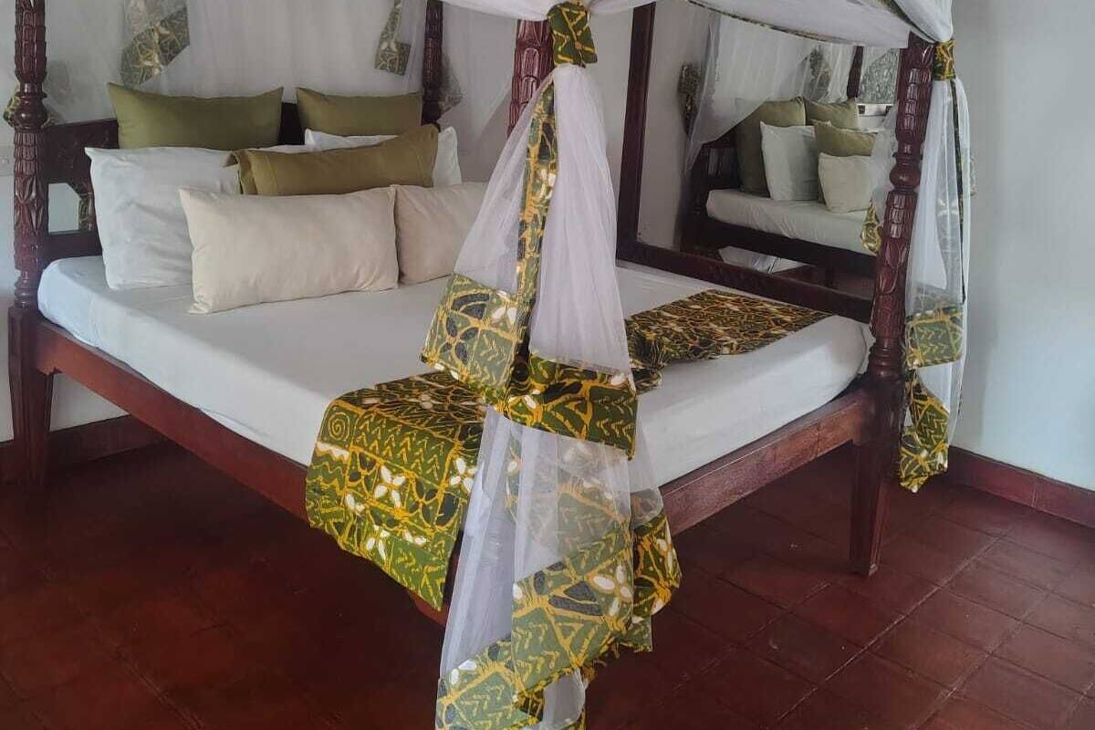 A four-poster canopy bed with a white and gold patterned bedspread, a wooden headboard, and a nightstand with a lamp.