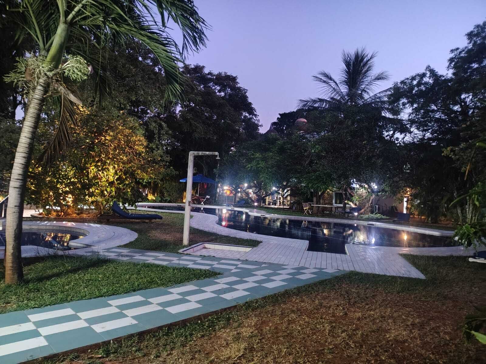 Un espacio al aire libre nocturno con piscina, un sendero a cuadros y palmeras.