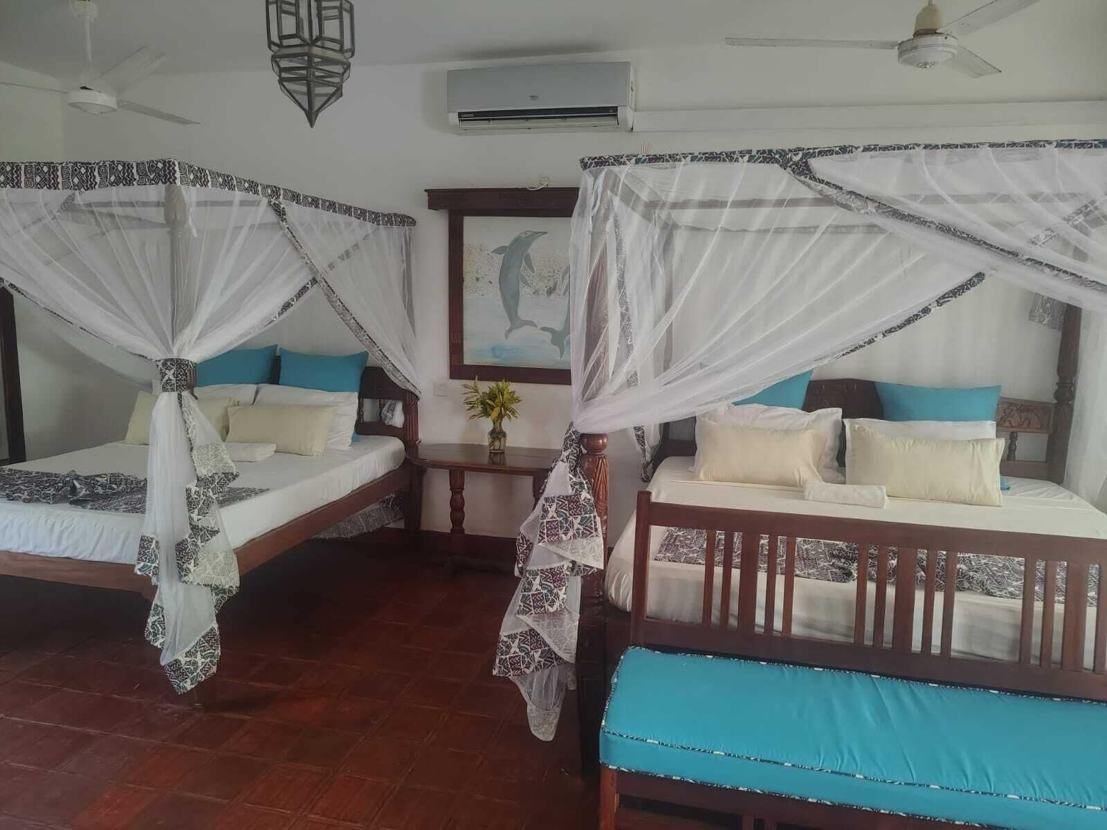 Two wooden beds with white bedding and blue cushions, each with a canopy. There is a small table with a vase of flowers between the beds.