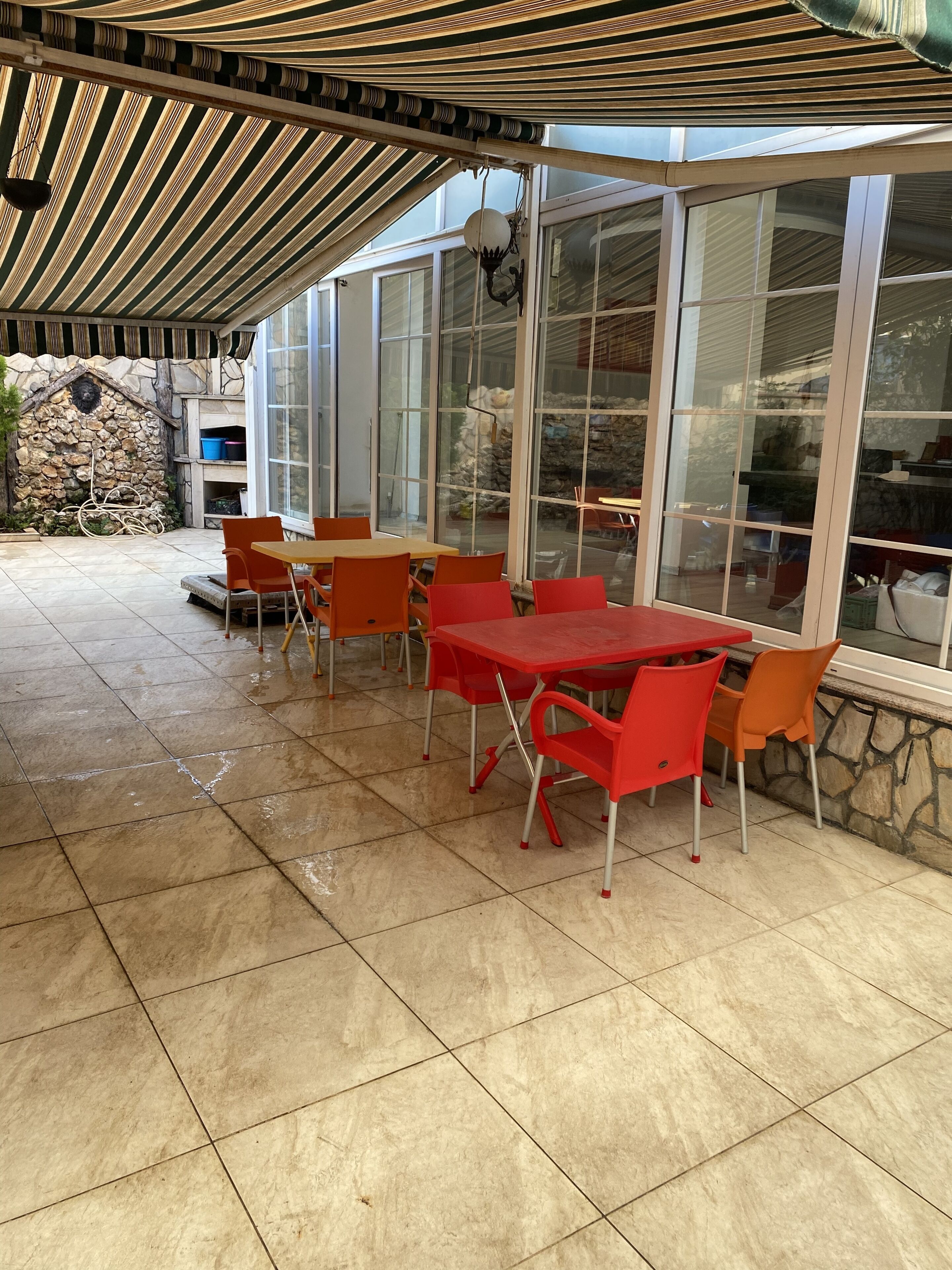 A covered patio area with tiled flooring, red and orange chairs, and tables. There are large windows providing views of the surrounding area.
