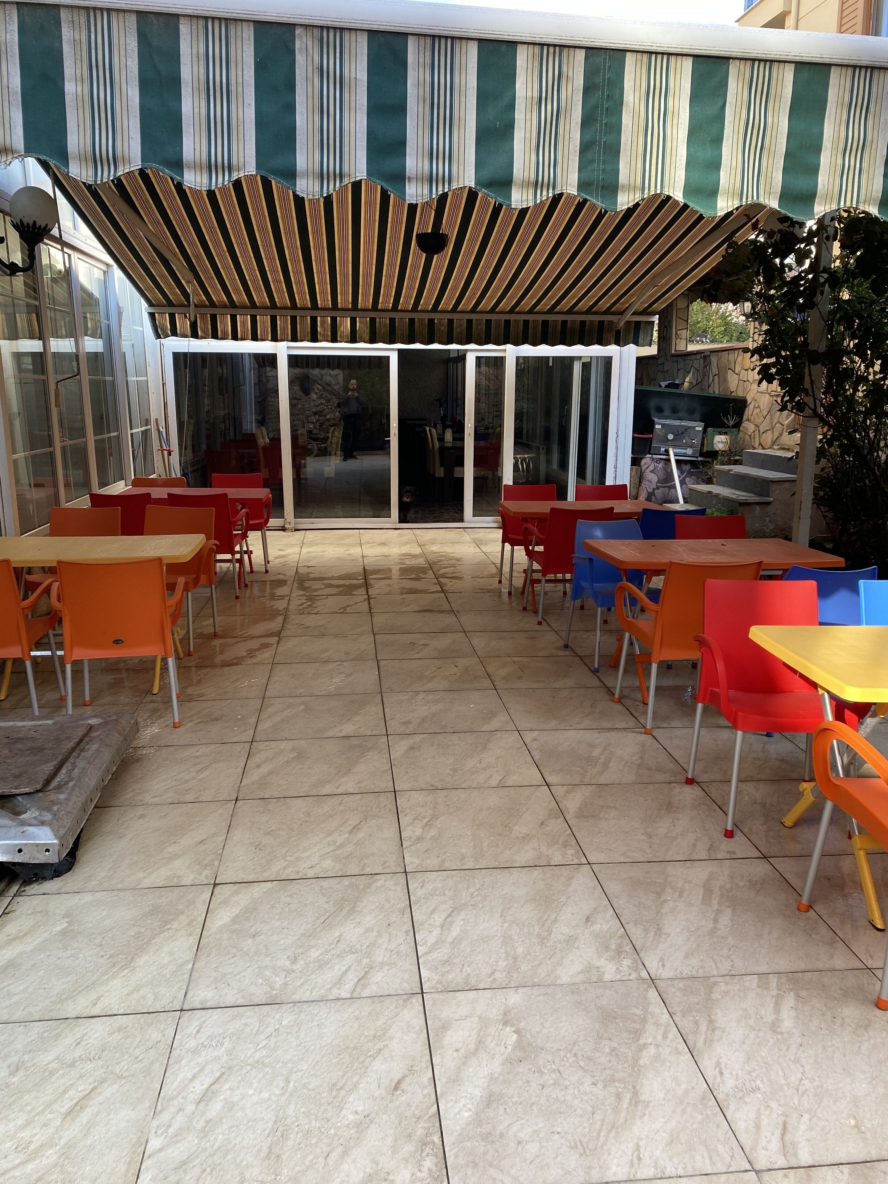 Un patio al aire libre con suelo de baldosas, sillas rojas y azules, y un toldo a rayas.
