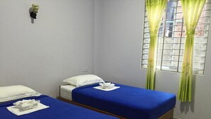 A room with two single beds, each with a blue bedspread and white pillows, a small wall-mounted nightstand, and a window with green curtains.