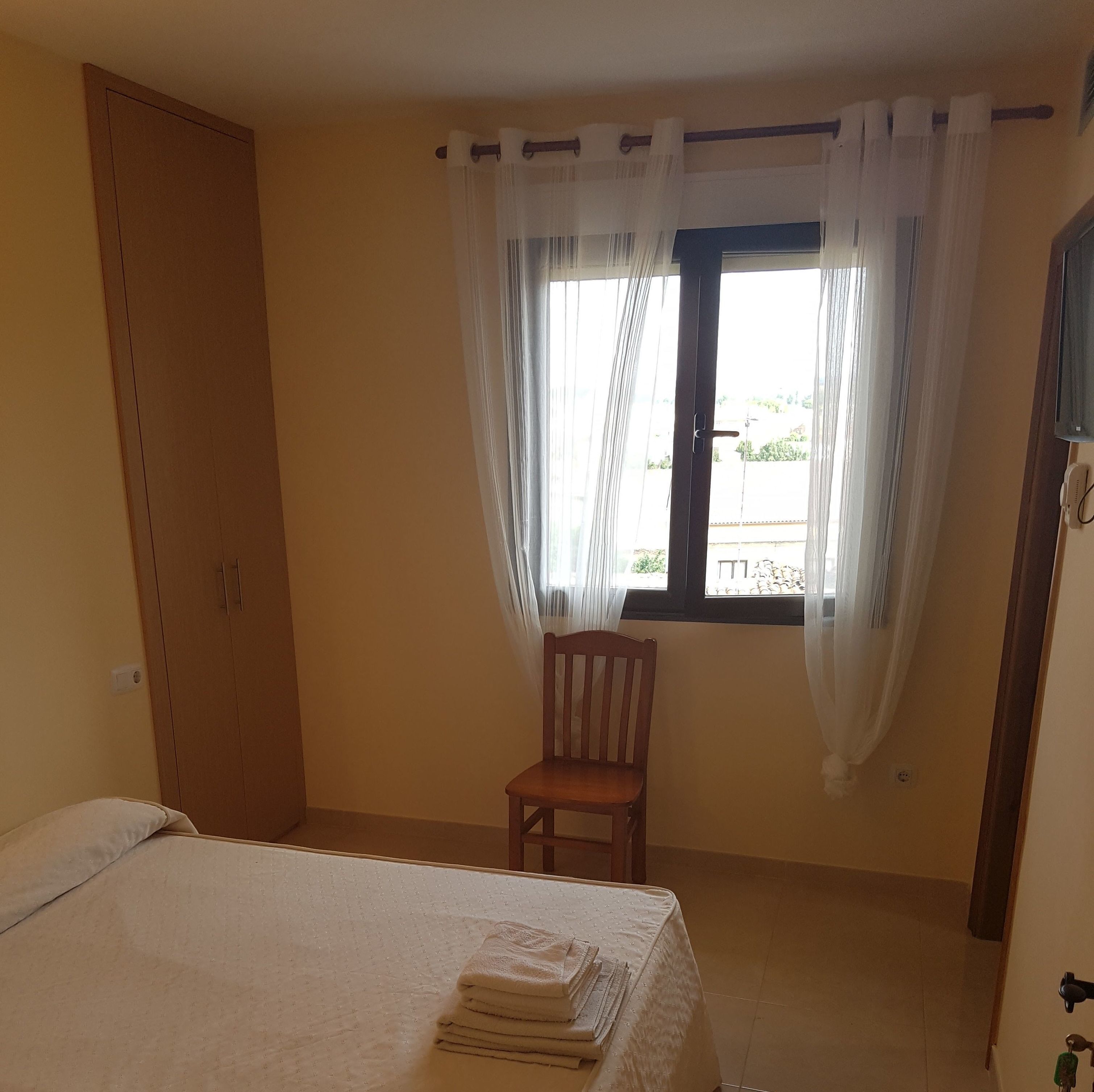 Una habitación con cama, un armario de madera, una silla y una ventana con cortinas traslúcidas.