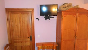 Quarto com porta de madeira, televisão fixada na parede, armário de madeira e uma pequena mesa com cadeira.
