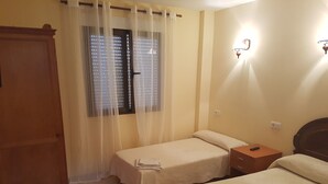 Quarto de hotel com duas camas, um guarda-roupa de madeira, uma janela com persianas e um candeeiro fixado na parede.