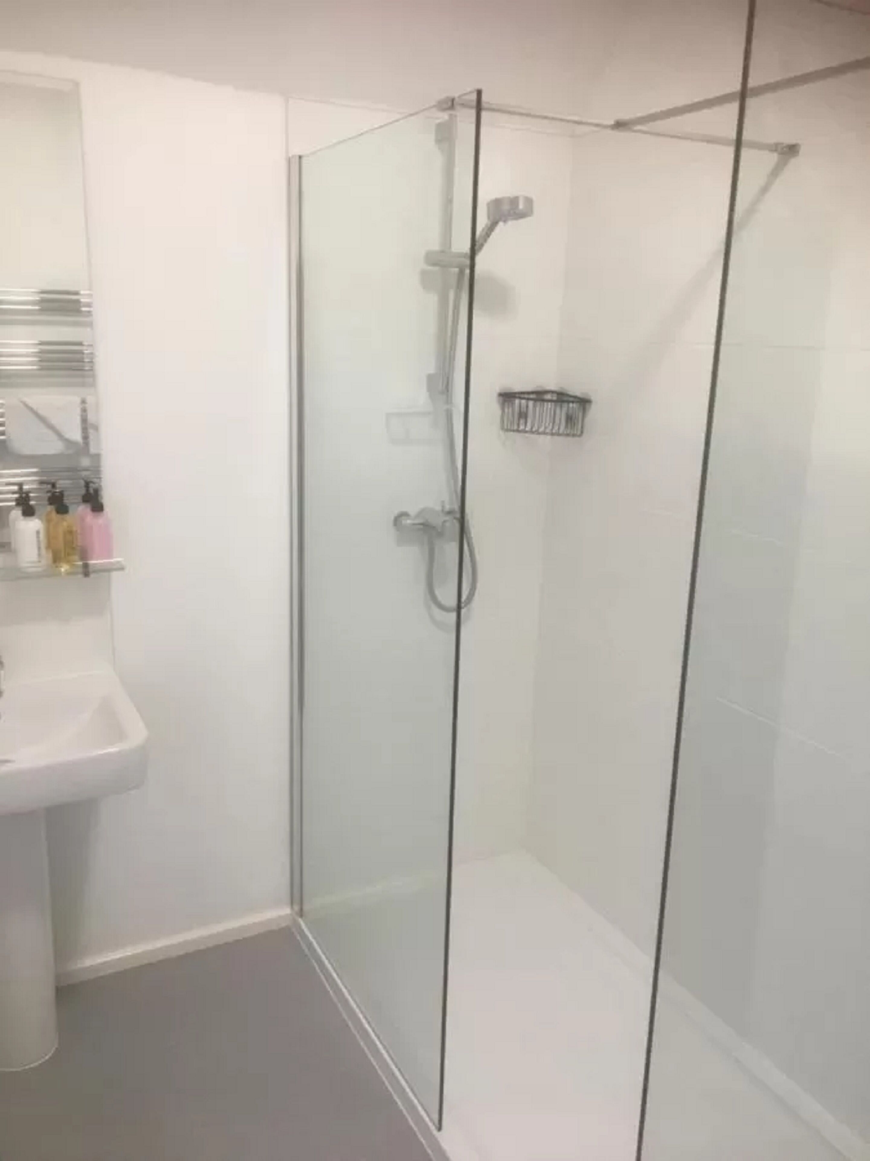 Une douche fermée par des parois en verre, dotée d’un pommeau de douche à main, d’une porte en verre et d’une petite étagère pour les articles de toilette.
