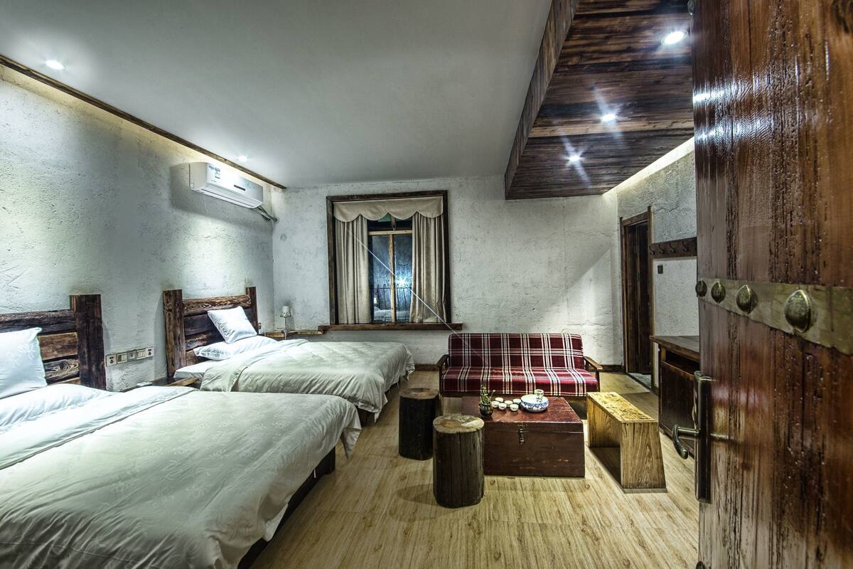 Een kamer met drie bedden, een houten bankje, een kleintje tafeltje met decoratieve objecten en een raam met gordijnen.