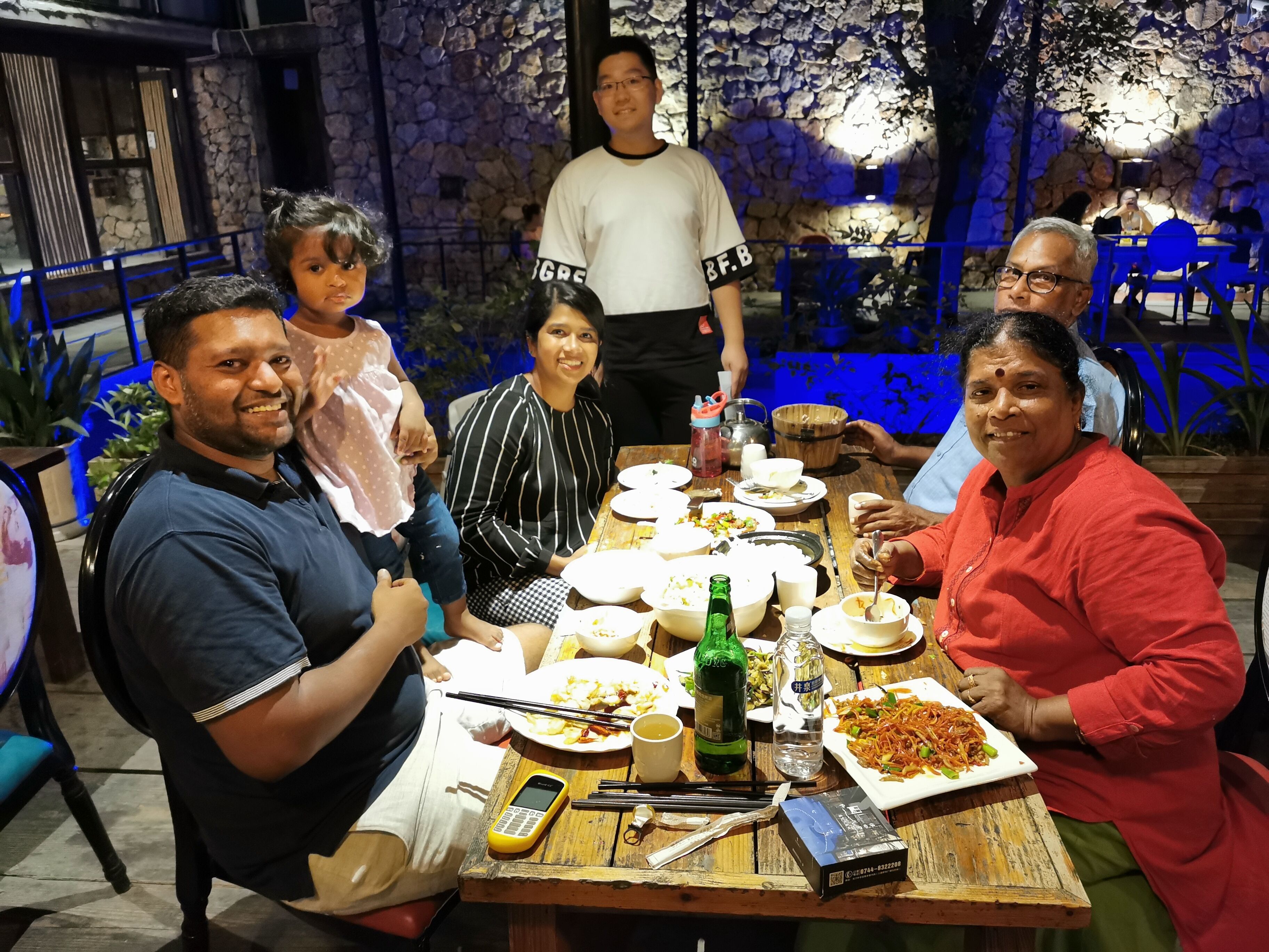 Un groupe de personnes est assis autour d’une table en bois, devant divers plats, une bouteille et un téléphone portable.