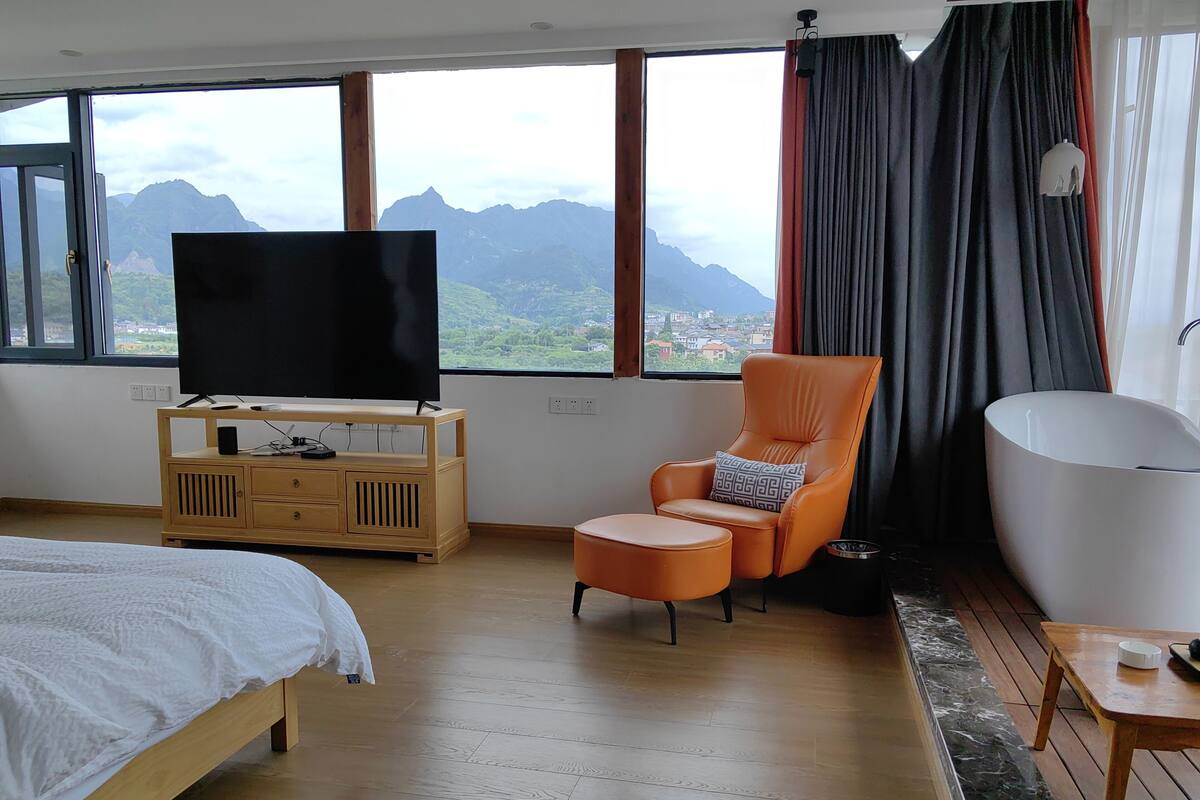 Een slaapkamer met een bed, een televisie op een houten standaard, een leren fauteuil, een witte badkuip en uitzicht op bergen door grote ramen.
