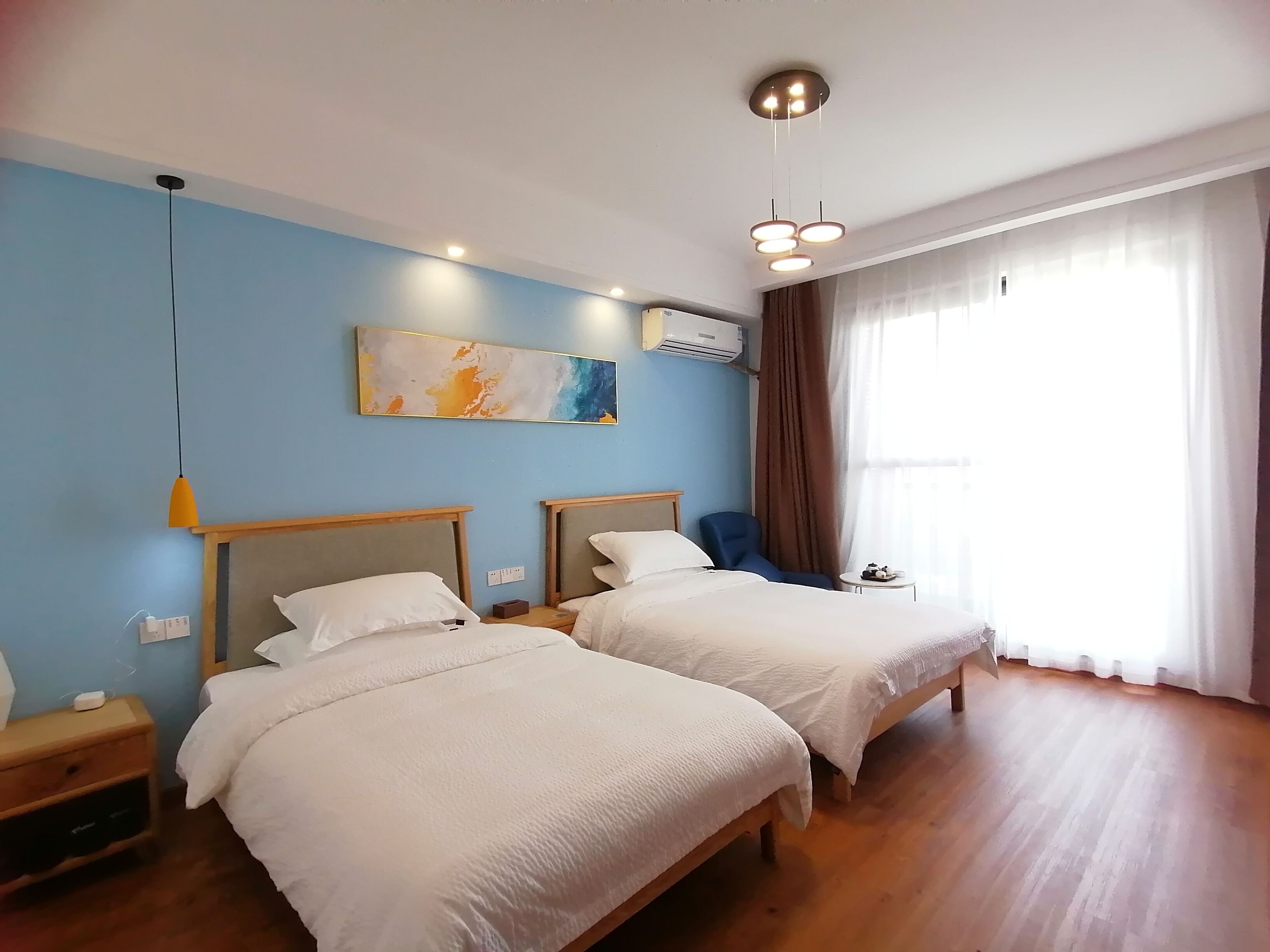 A hotel room with two beds, a blue wall, a ceiling light, and a window with curtains.