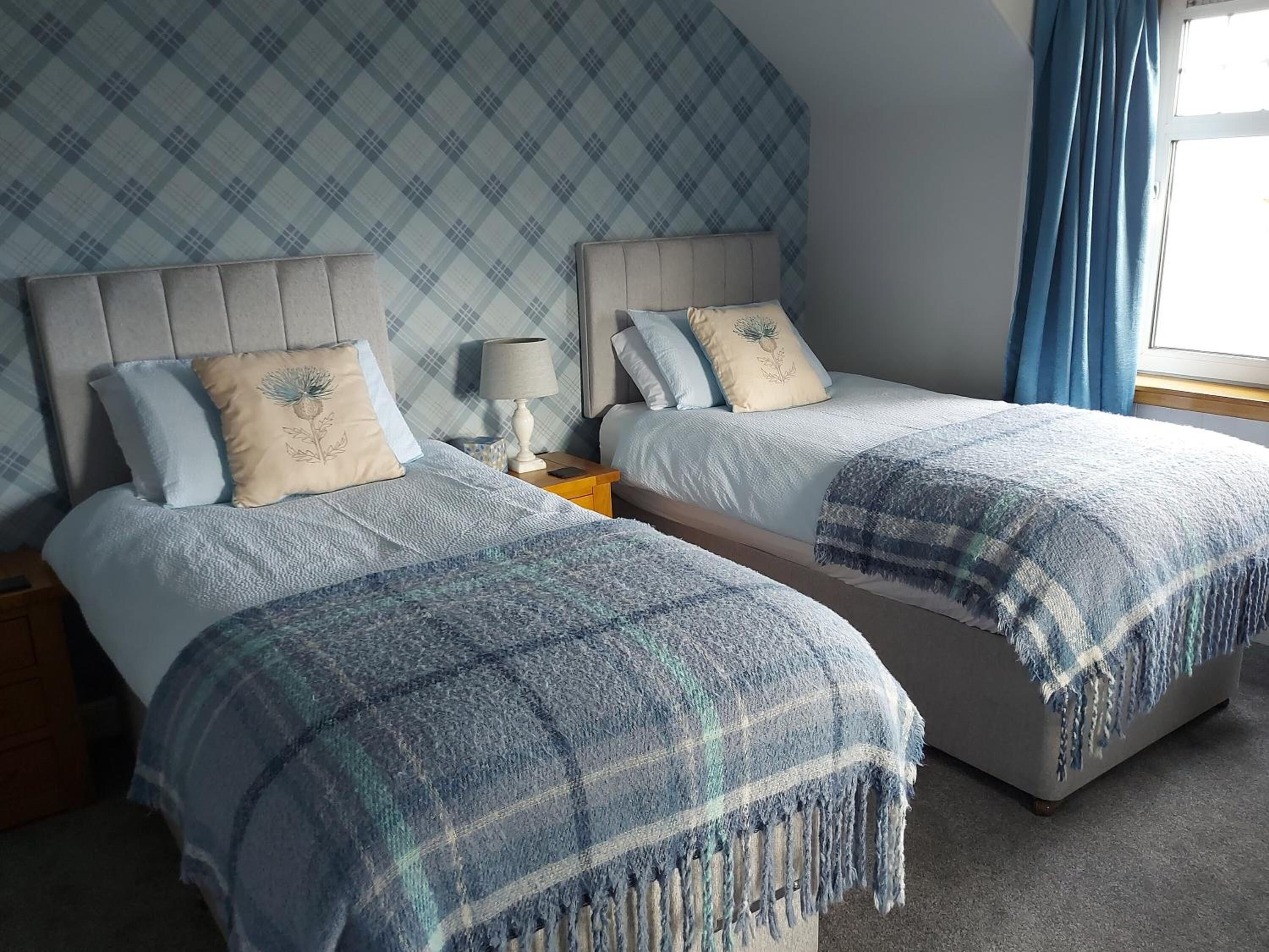 A twin bedroom with two beds, a nightstand, a lamp, and a window with curtains.