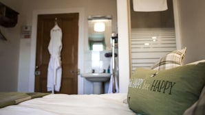 A hotel room with a bed, a green pillow with "HAPPY bee HAPPY" text, a robe hanging on a door, and a bathroom with a sink and mirror. - Fir Tree B&B at Galvelbeg House (Crieff)