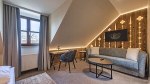 Ein gemütliches, modernes Schlafzimmer im Loft-Stil mit schrägem Dach, großem Fenster mit Vorhängen, einer Couch, einem Couchtisch und einem an einer Holzwand montierten Fernseher.