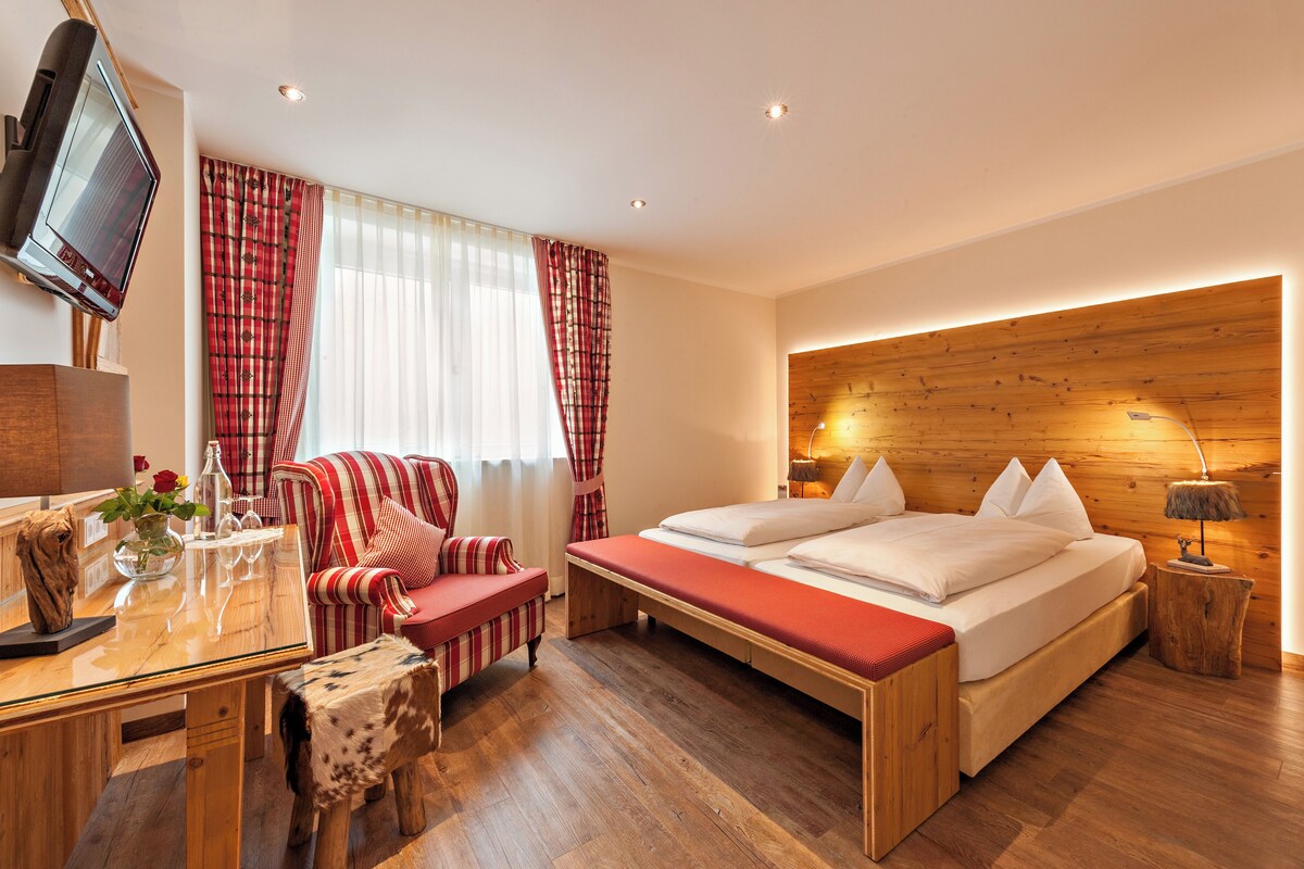 A modern hotel room with a bed, a TV, a desk with a vase of flowers, and a red-striped armchair.
