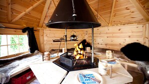 A wooden cabin interior with a central wood-burning stove, a dining table with plates and cups, and a fur-covered bench. - Hotel Schäfflerwirt (Aschheim)