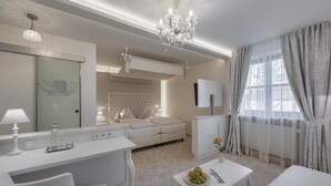 Ein modernes Hotelzimmer mit weißer Farbgestaltung, einer Kristallleuchter, einem Bett mit gestepptem Kopfteil, einem Schreibtisch mit Lampe sowie einem Esstisch mit Früchten.