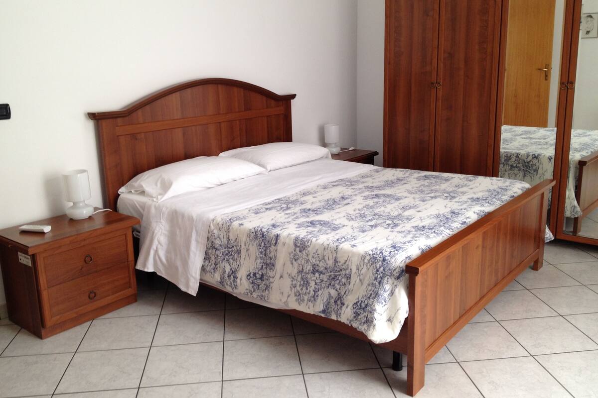 A wooden bed with a floral bedspread, a nightstand with a lamp, and a wardrobe in a hotel room.