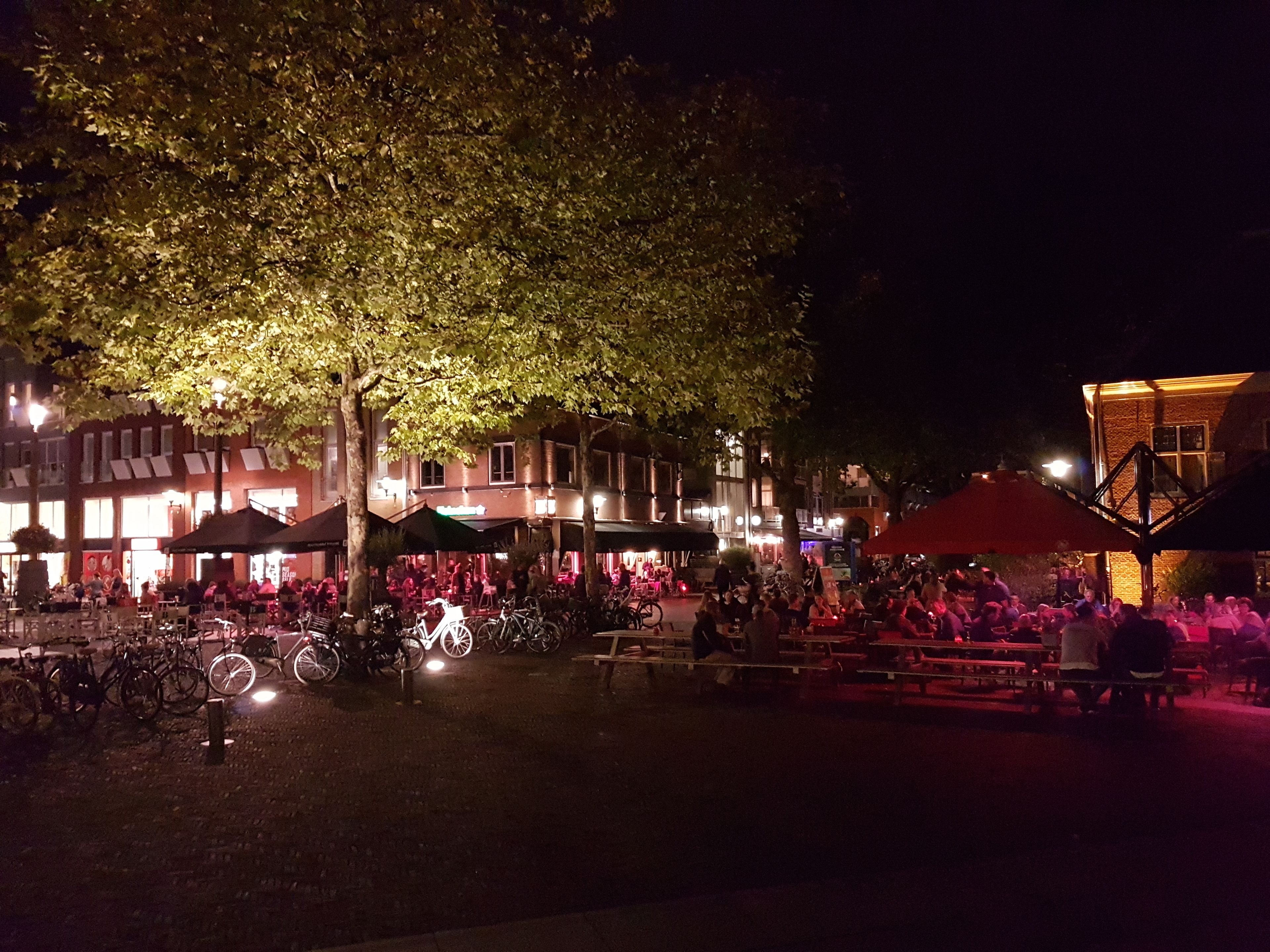 An outdoor evening setting with tables and chairs under trees, surrounded by buildings with illuminated windows.