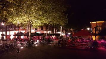An outdoor evening setting with tables and chairs under trees, surrounded by buildings with illuminated windows.