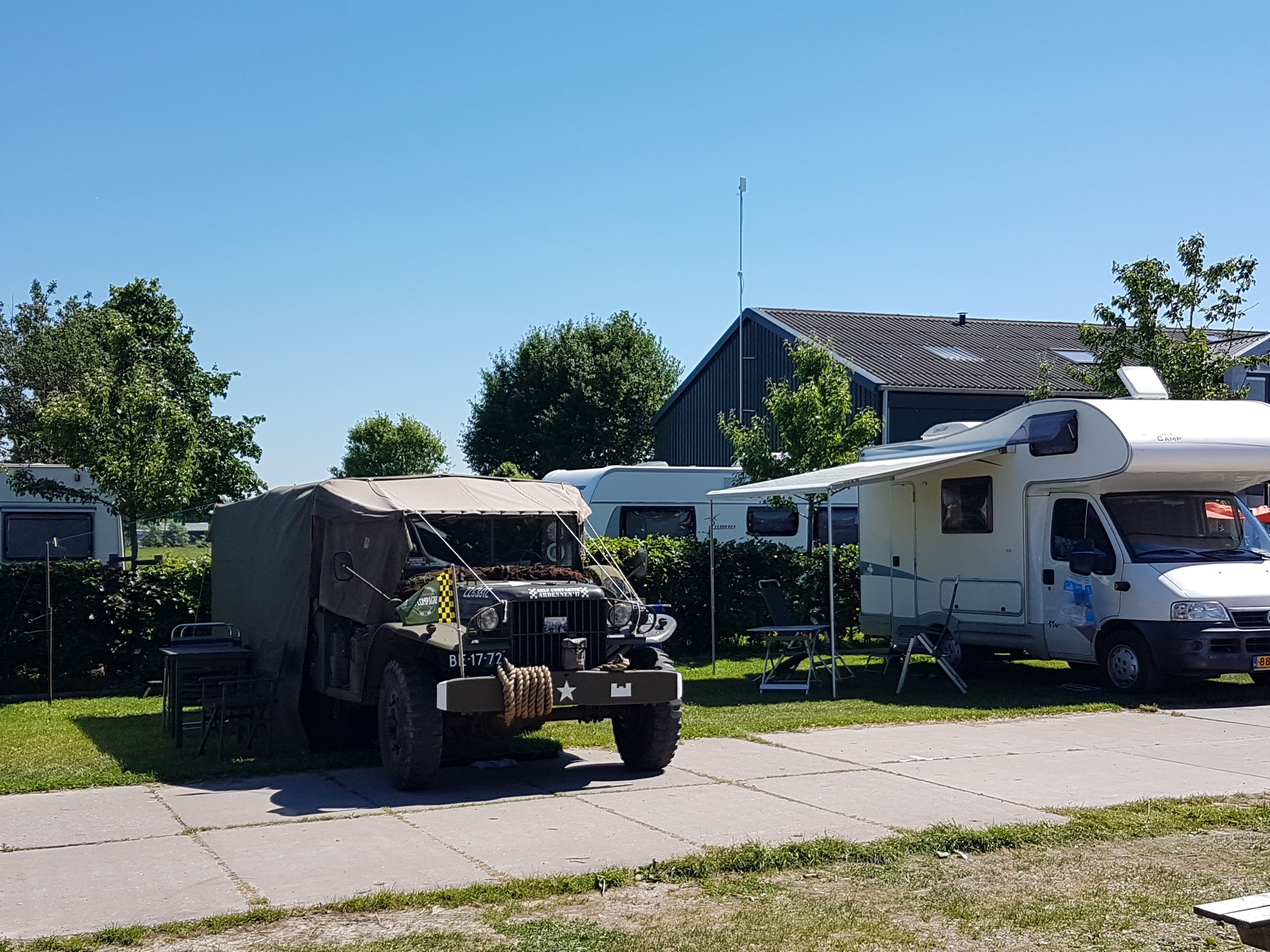 A military-style vehicle parked in a residential area with a large RV and a house in the background.