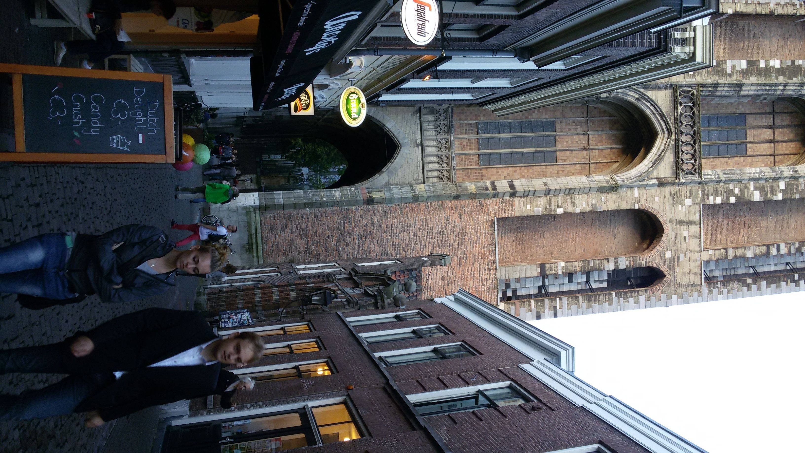 A street view with a brick building, a signboard with prices, and a few people walking.