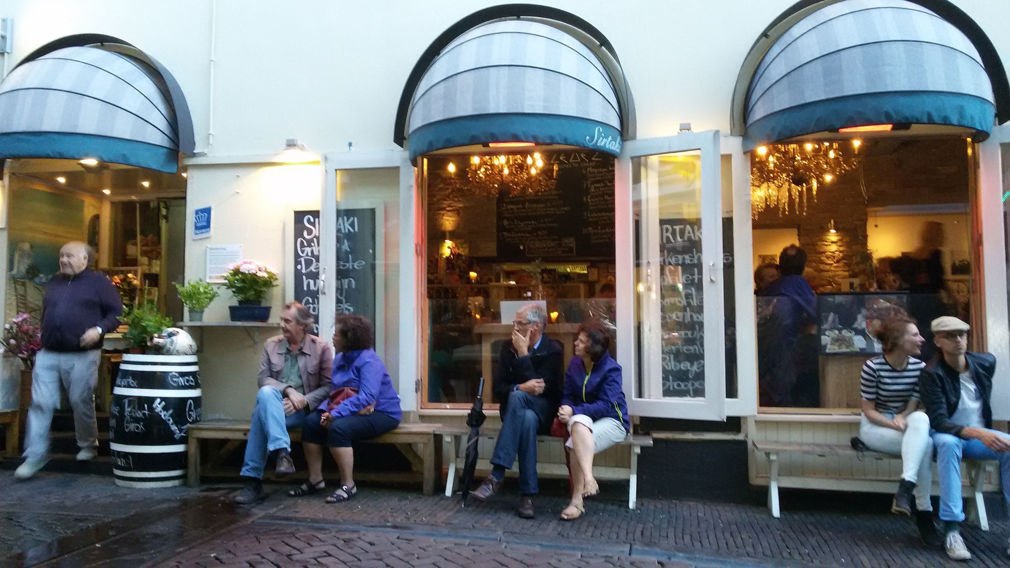A group of people sitting on outdoor benches outside a building with arched windows and a sign that reads "Sister".