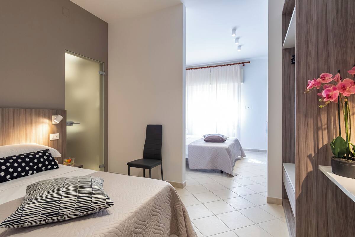 Une chambre d’hôtel moderne comprenant un lit, une chaise, une petite table, une table de chevet, une plante en pot et une vue sur la chambre voisine.