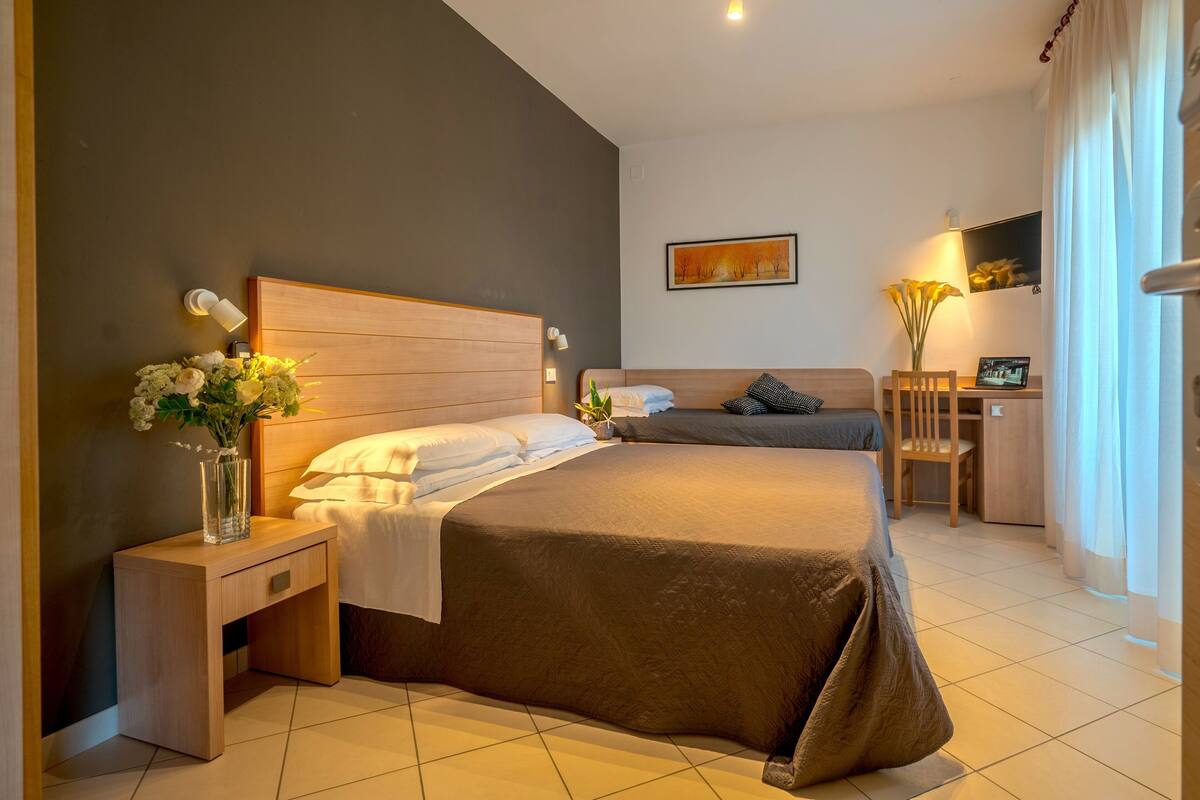 Une chambre d’hôtel moderne avec un grand lit, des tables de chevet, une petite table, une chaise, un bureau et un vase avec des fleurs.
