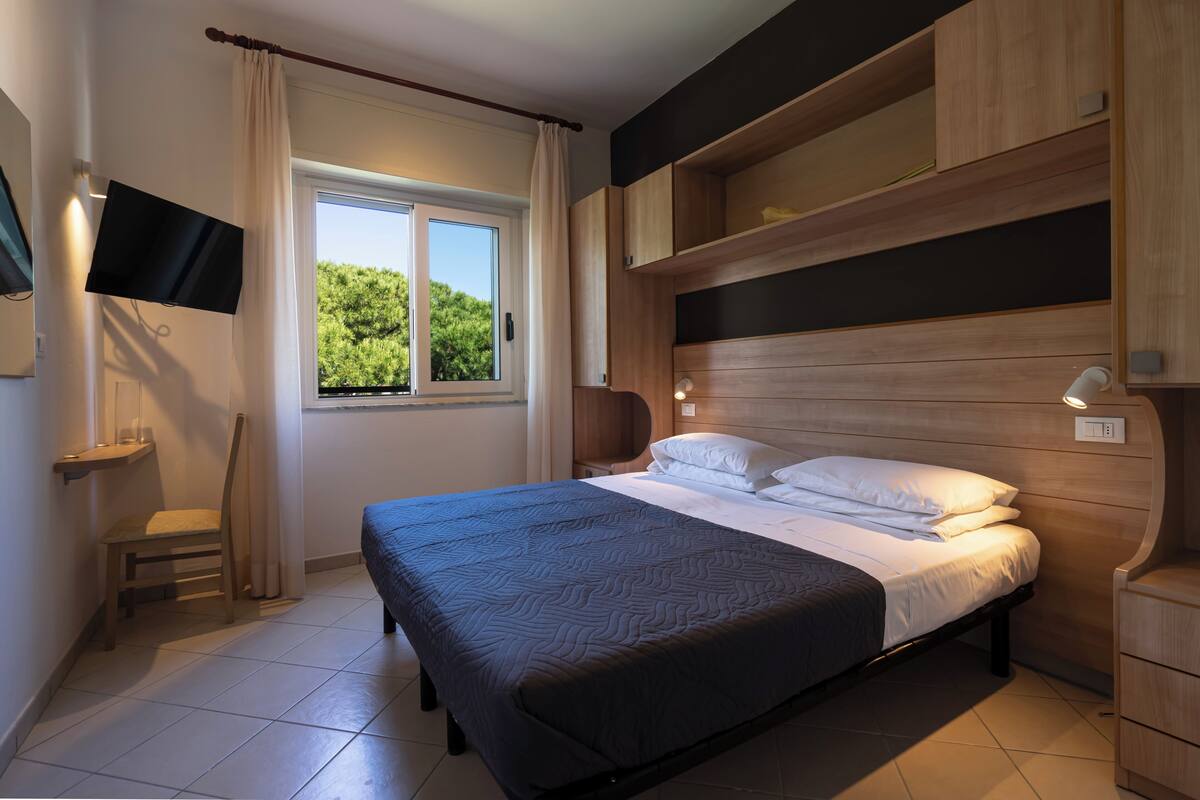 Une chambre à coucher comprenant un lit, un téléviseur, une fenêtre avec des rideaux, une chaise et des étagères en bois intégrées.