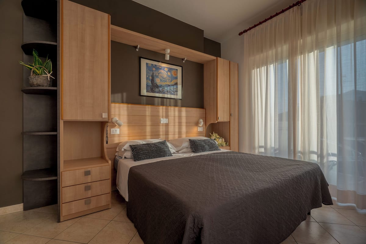Une chambre avec un grand lit, des étagères intégrées, un tableau au-dessus du lit et une fenêtre avec des rideaux légers.