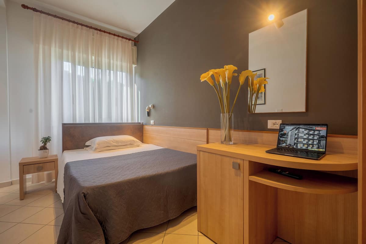Une chambre d’hôtel avec un lit, une table de chevet, une lampe, un vase avec des fleurs et un ordinateur portable posé sur un bureau en bois.
