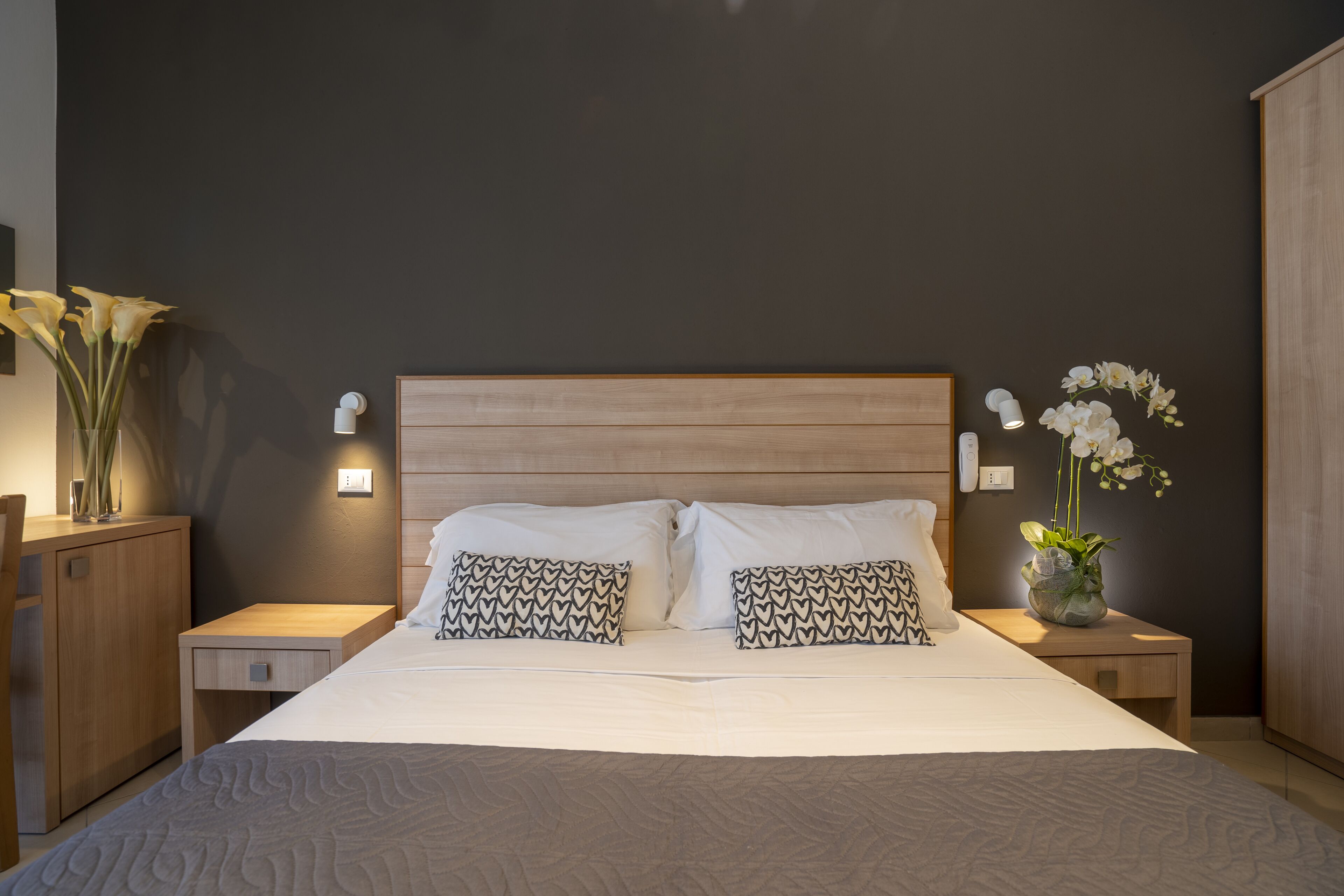 Ein ordentlich bezogenes Bett mit gemusterten Kissen, ein Nachttisch mit Blumenvase und eine an der Wand befestigte Leuchte.