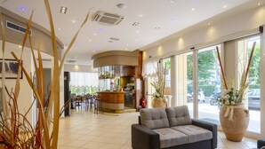 A modern hotel lobby with a seating area, a reception desk, and large windows offering a view of the street. - Hotel Mocambo (Riccione)
