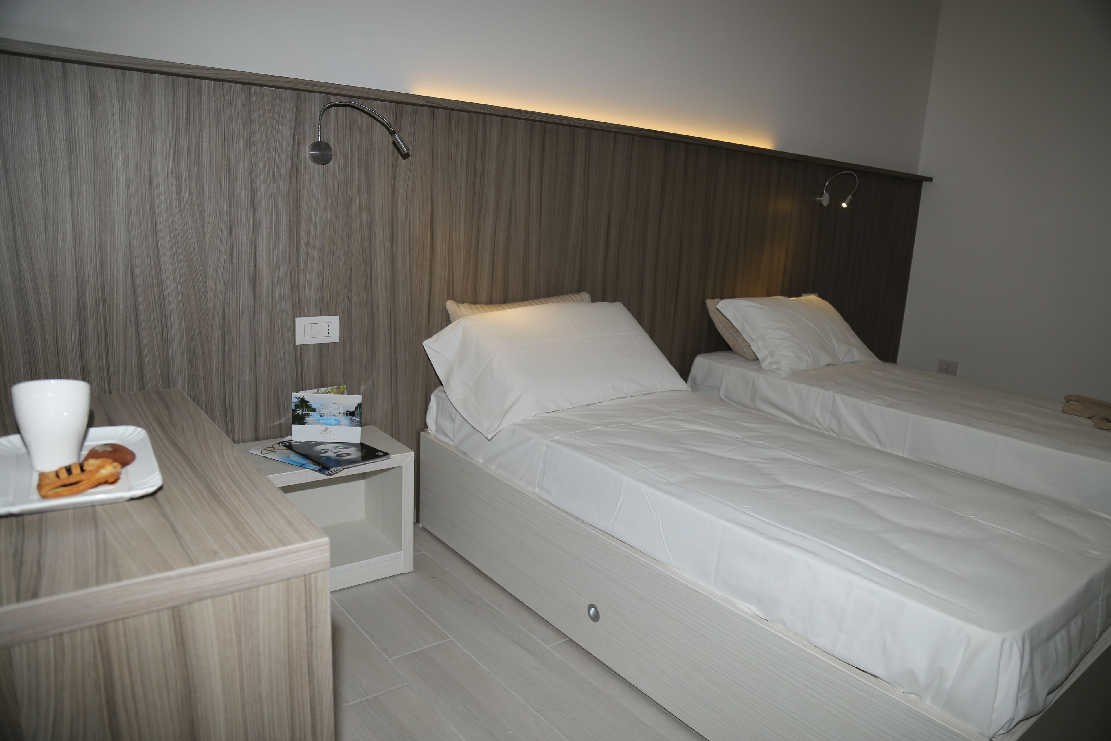 A hotel room with two single beds, a bedside table, a nightstand, and a small desk with a cup and a plate.