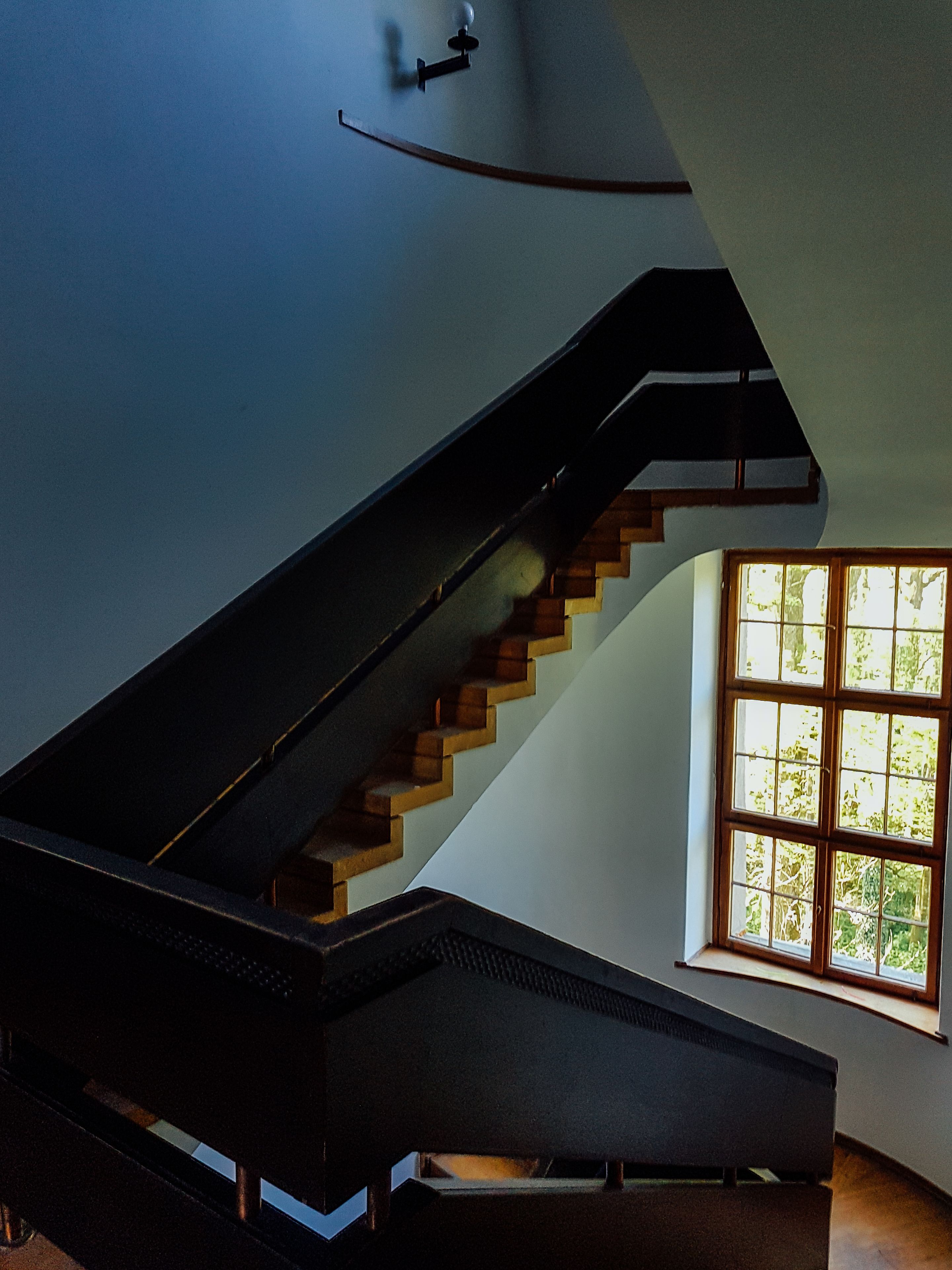 Un escalier moderne avec une rampe noire et des marches en bois, menant à un palier doté d’une fenêtre qui offre une vue sur la verdure environnante.