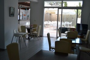 A dining area with white chairs and tables, a television, and a view of an outdoor patio with white chairs and umbrellas. - Ifigenia Hotel (Skiathos)