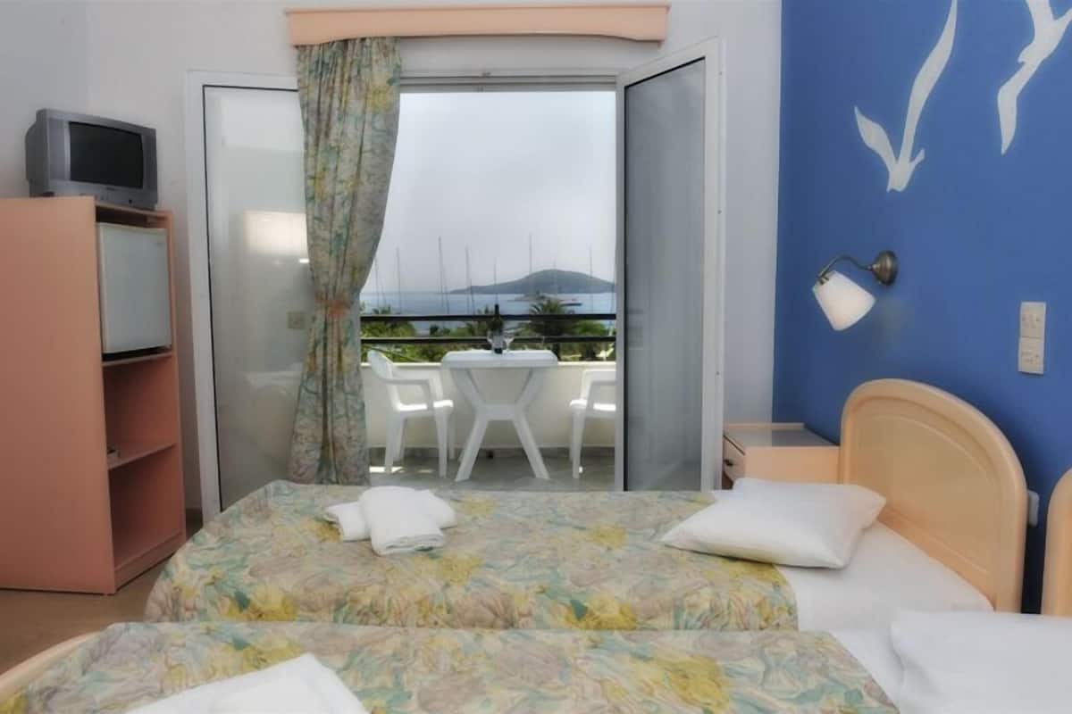 Quarto de hotel com duas camas, televisão, varanda com mesa e cadeiras, e vista para o mar.