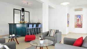 A modern living room with a grey sofa, red ottoman, and glass coffee table. There is a kitchen area with a blue cabinet and a bar counter. - Ifigenia Hotel (Skiathos)