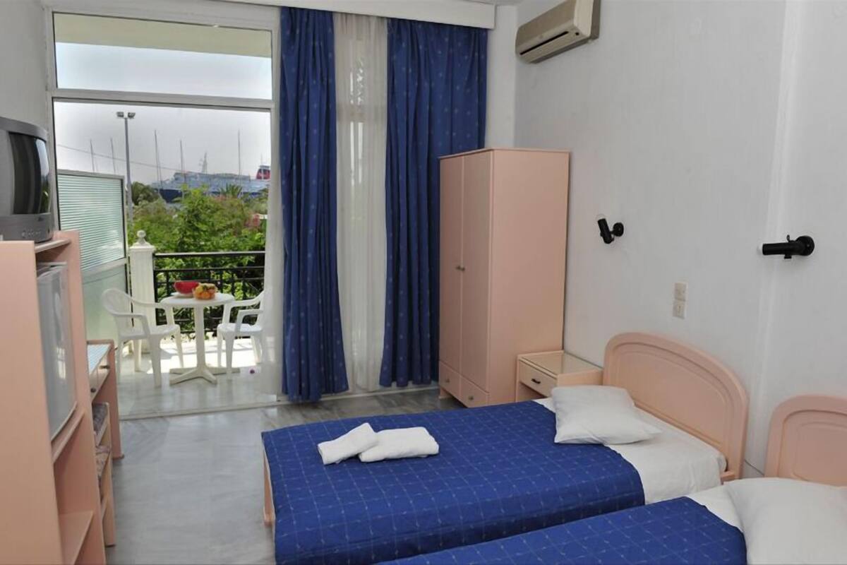 Quarto de hotel com duas camas, um guarda-roupa, uma escrivaninha com cadeira e varanda com mesa e cadeiras.