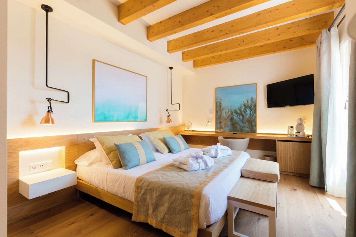 A modern hotel room with a wooden ceiling, a bed with a beige blanket, a desk with a chair, and artwork on the walls.