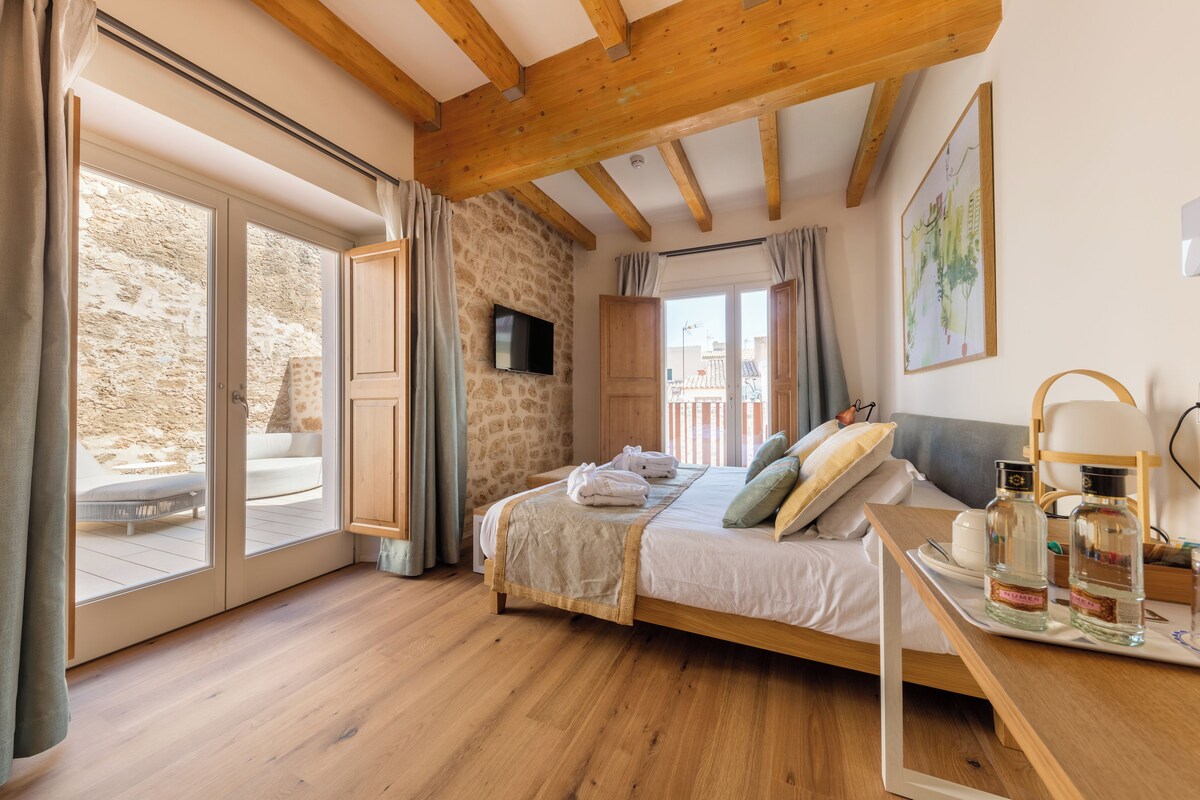 A bedroom with a wooden ceiling, a stone wall, a bed with pillows, a TV, and a balcony with a table and chairs.