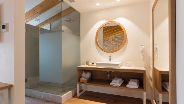 Une salle de bain moderne avec une douche à l’italienne en verre, un meuble-lavabo en bois avec un miroir rond et une étagère intégrée où sont rangées les serviettes.
