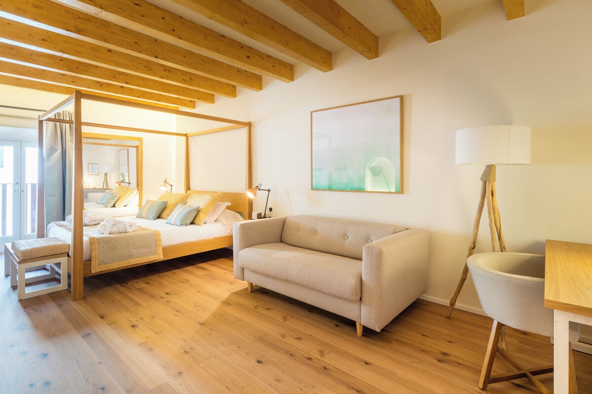 A modern bedroom with a wooden ceiling, a bed with a headboard, a beige sofa, a wooden table, and a chair.