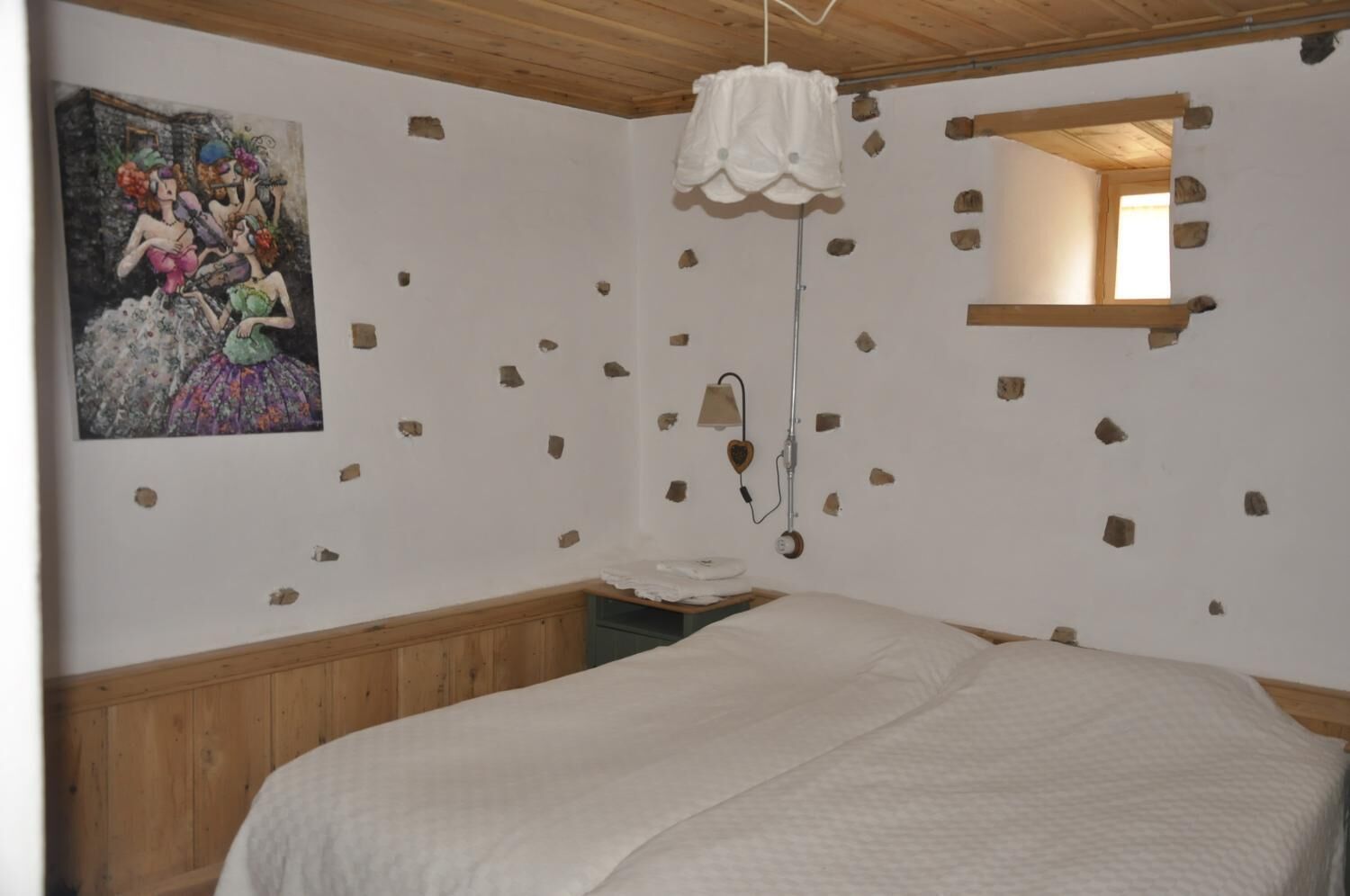 Een slaapkamer met een houten hoofdbord, een witte spreidek en een hanglamp, met een lamp in de vorm van een hart, en een schilderij aan de muur.