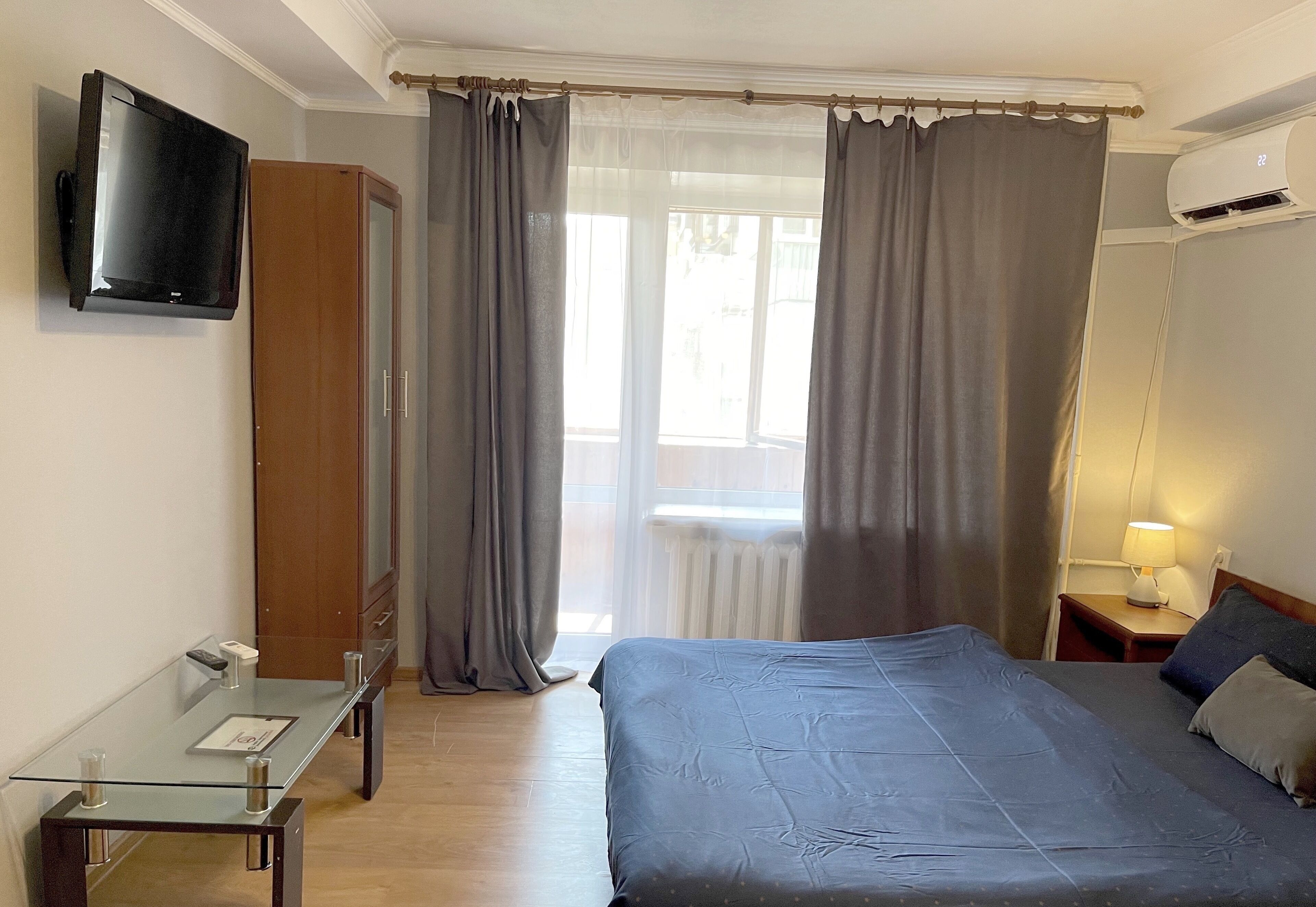 A bedroom with a bed, a TV mounted on the wall, a desk with a chair, a wardrobe, and a window with curtains.
