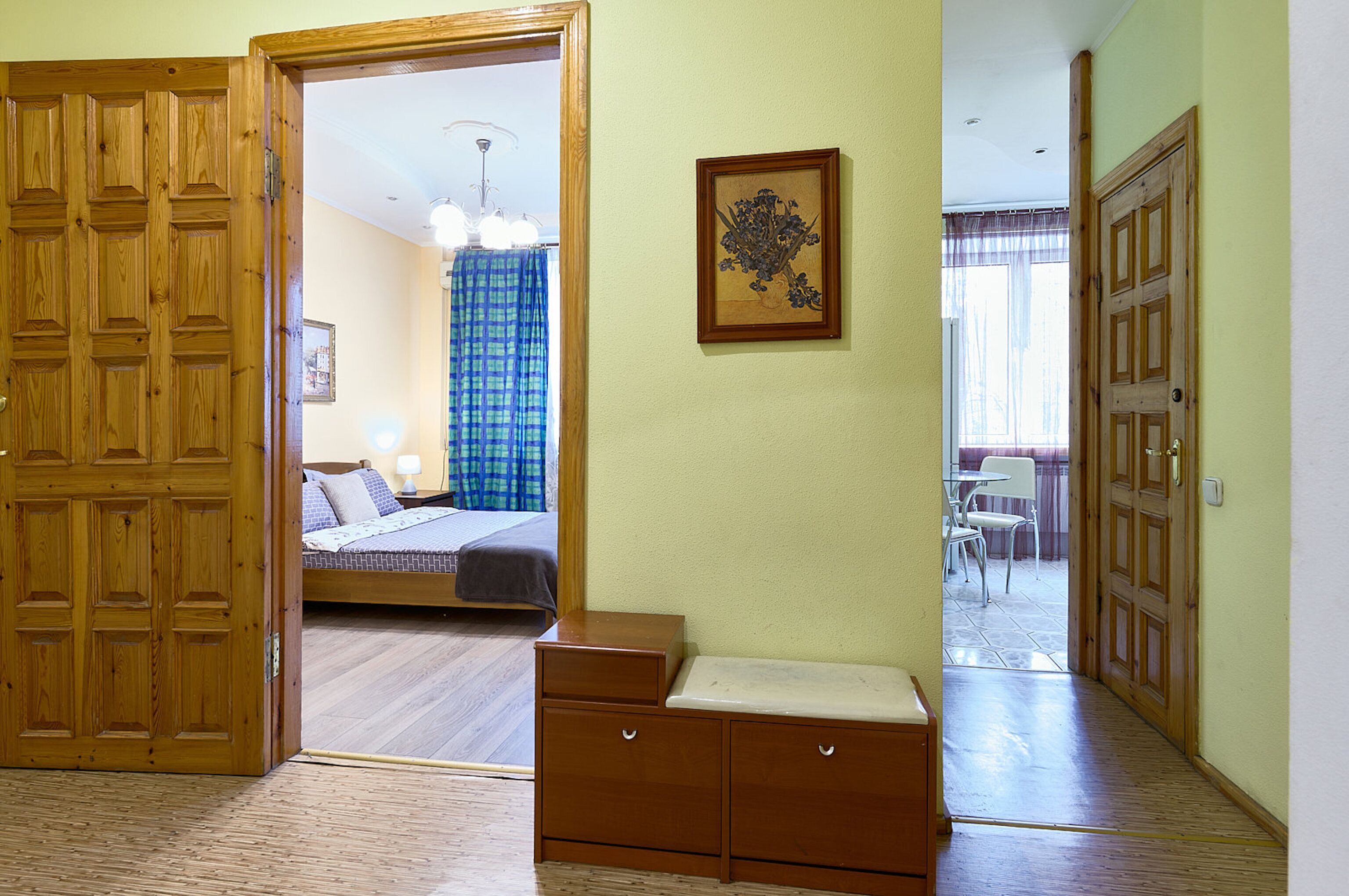 A room with a wooden wardrobe, a bedside table, a bed with a blue and white checkered blanket, and a framed picture on the wall.