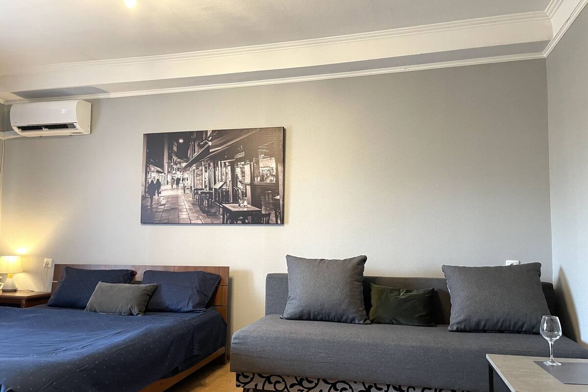 A hotel room with two beds, a sofa, a table with a glass, and a wall-mounted air conditioner.